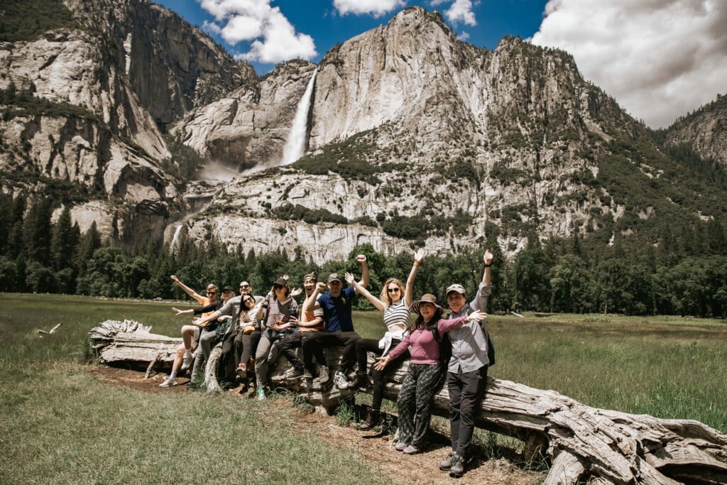 a group of smiling people sitting at a fallen tree trunk with a view of yosemite falls