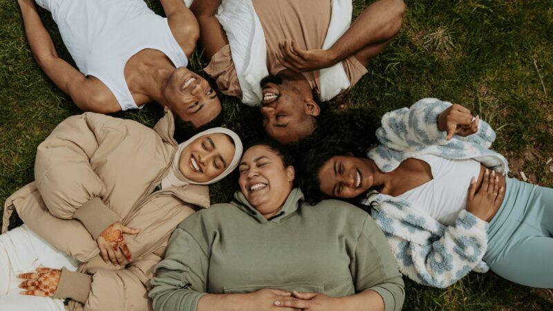 A diverse group of five friends lying on the grass in a circle, laughing and smiling, dressed in cozy clothing.