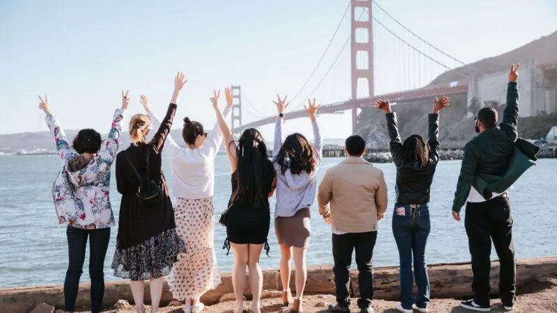 A group of eight people stands with their backs to the camera, raising their hands in the air while facing the Golden Gate Bridge in San Francisco