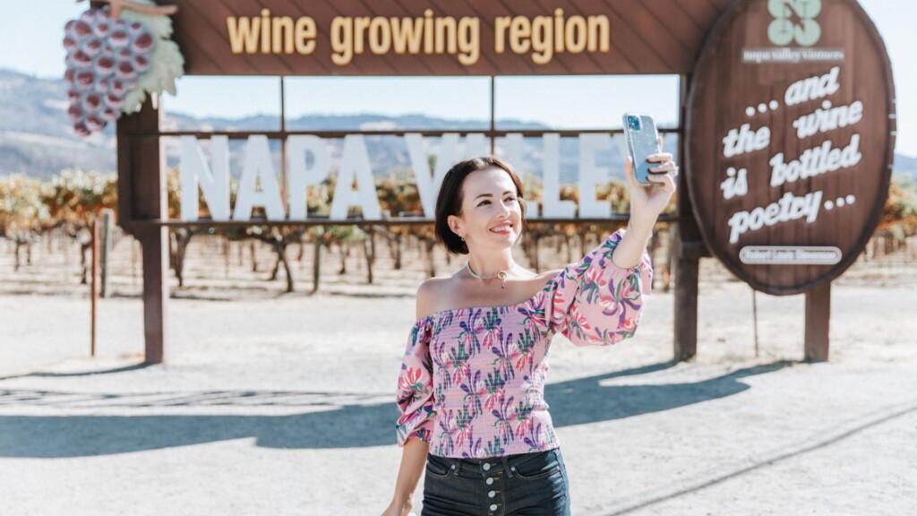 A woman in a floral off-shoulder top takes a selfie in front of a large wooden sign that reads 'NAPA VALLEY'