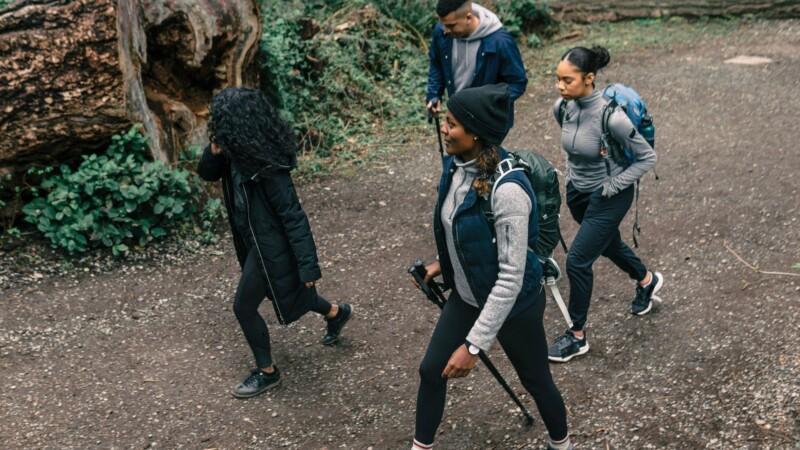 A group of four people, dressed in warm clothing and carrying backpacks, are hiking on a dirt trail in a wooded area.
