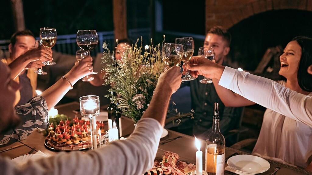 A group of friends gathers around a candlelit dinner table, raising their wine glasses in a toast.
