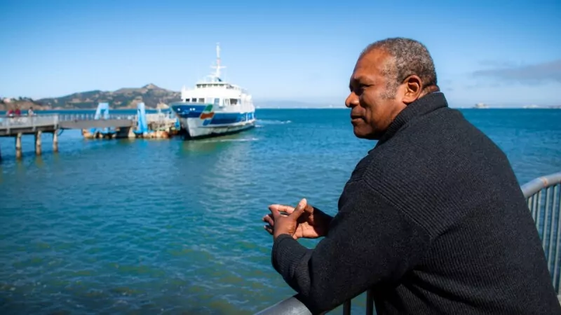 A man wearing a dark sweater is leaning on a metal railing, gazing thoughtfully at the water.
