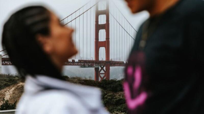 A blurred couple is standing in the foreground, facing each other, with the Golden Gate Bridge in sharp focus in the background.