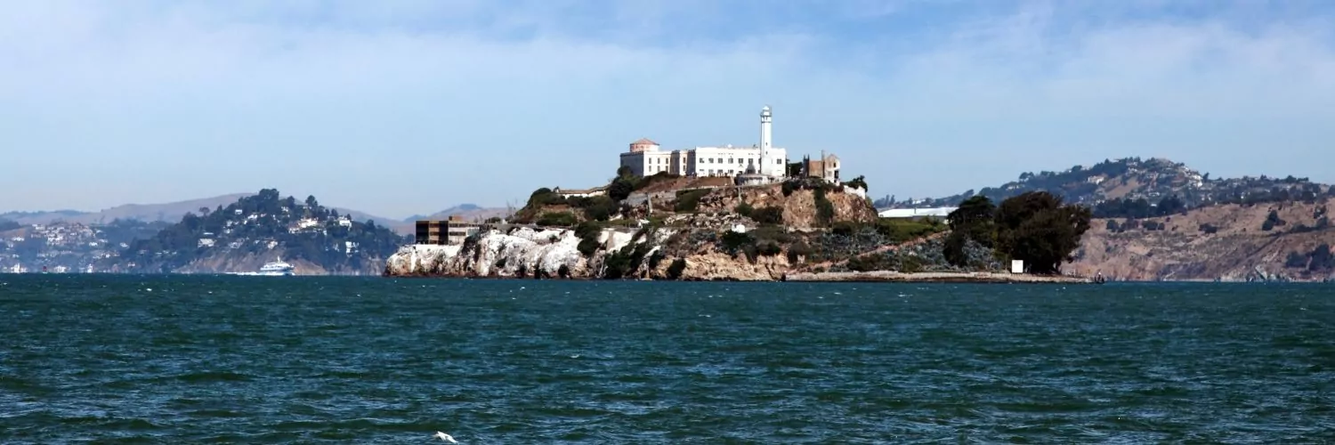 Alcatraz Island in San Francisco Bay, featuring the historic federal prison, a tall water tower, and rugged cliffs surrounded by blue waters under a clear sky