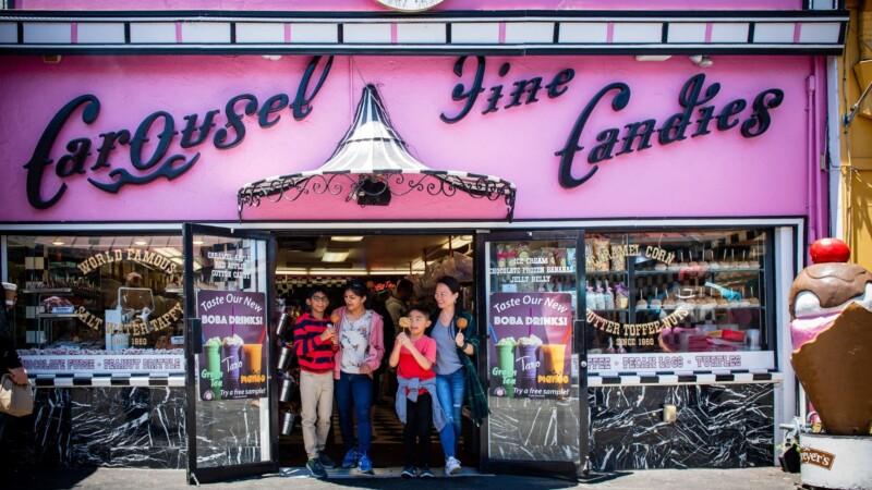 Family enjoying treats outside Carousel Fine Candies shop with pink storefront and vintage signage in a lively boardwalk setting