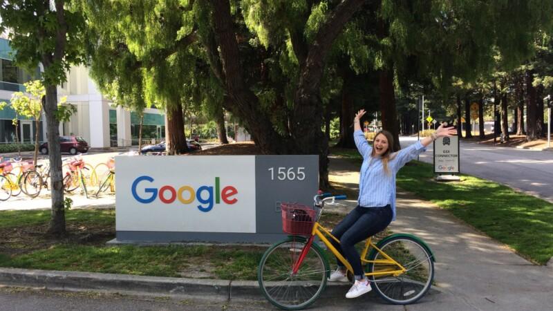 Woman smiling and posing on a colorful bike in front of the Google headquarters sign at 1565, surrounded by trees and campus buildings