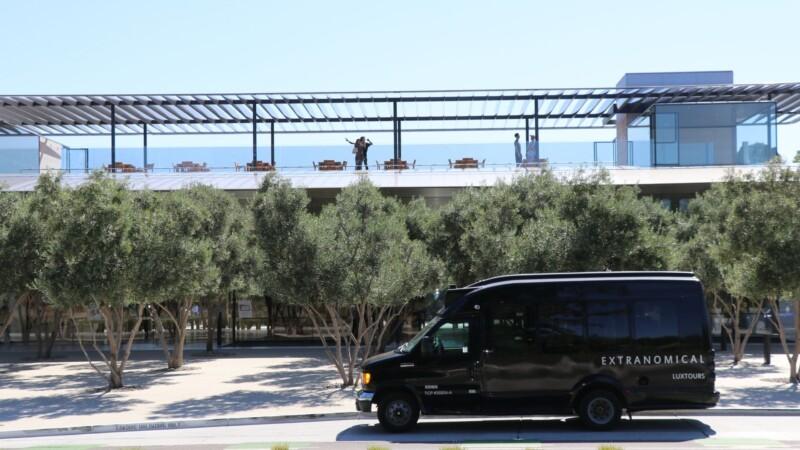 A sleek Extranomical tour van parked in front of a modern, glass-walled building with rooftop seating and visitors enjoying the view.