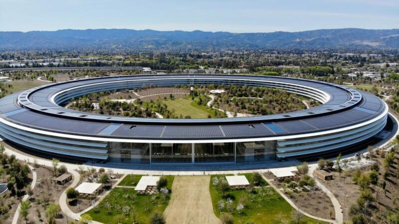 Aerial view of Apple Park, the circular headquarters of Apple Inc. in Cupertino, California, surrounded by landscaped greenery and mountains in the distance under a clear blue sky