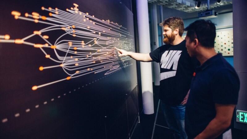 Two men discuss a complex data visualization displayed on a large screen.