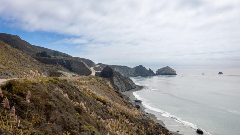 Scenic view of the California coastline with a winding highway, rocky cliffs, and the Pacific Ocean under a partly cloudy sky
