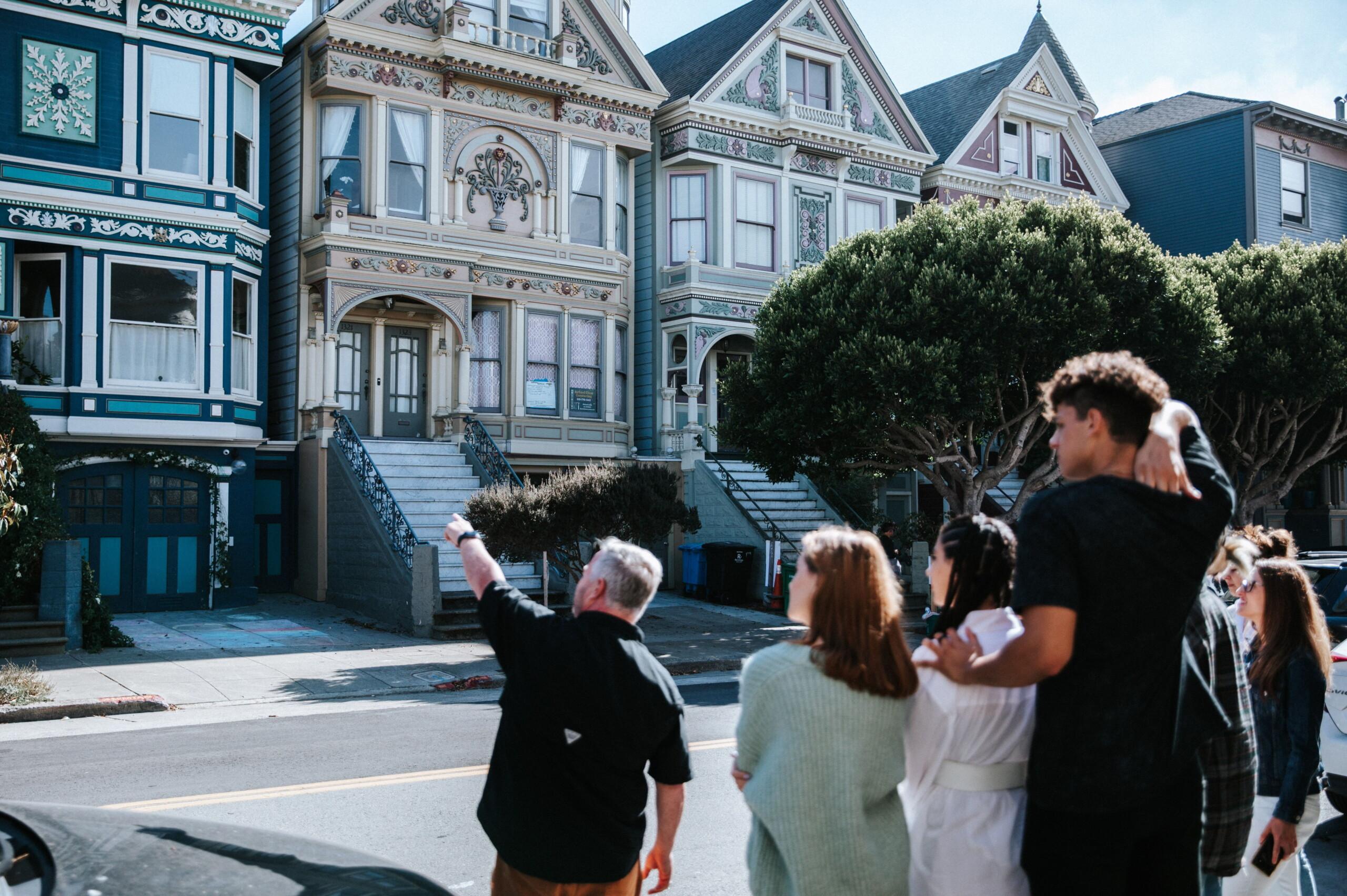 A group of people standing on the sidewalk in front of colorful Victorian-style houses, with some pointing and taking photos. A group of people standing on the sidewalk in front of colorful Victorian-style houses, with some pointing and taking photos.